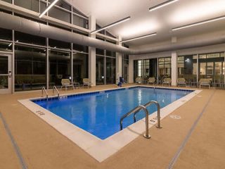 A well-lit indoor hotel pool at night with clear blue water, surrounded by white chairs and expansive windows